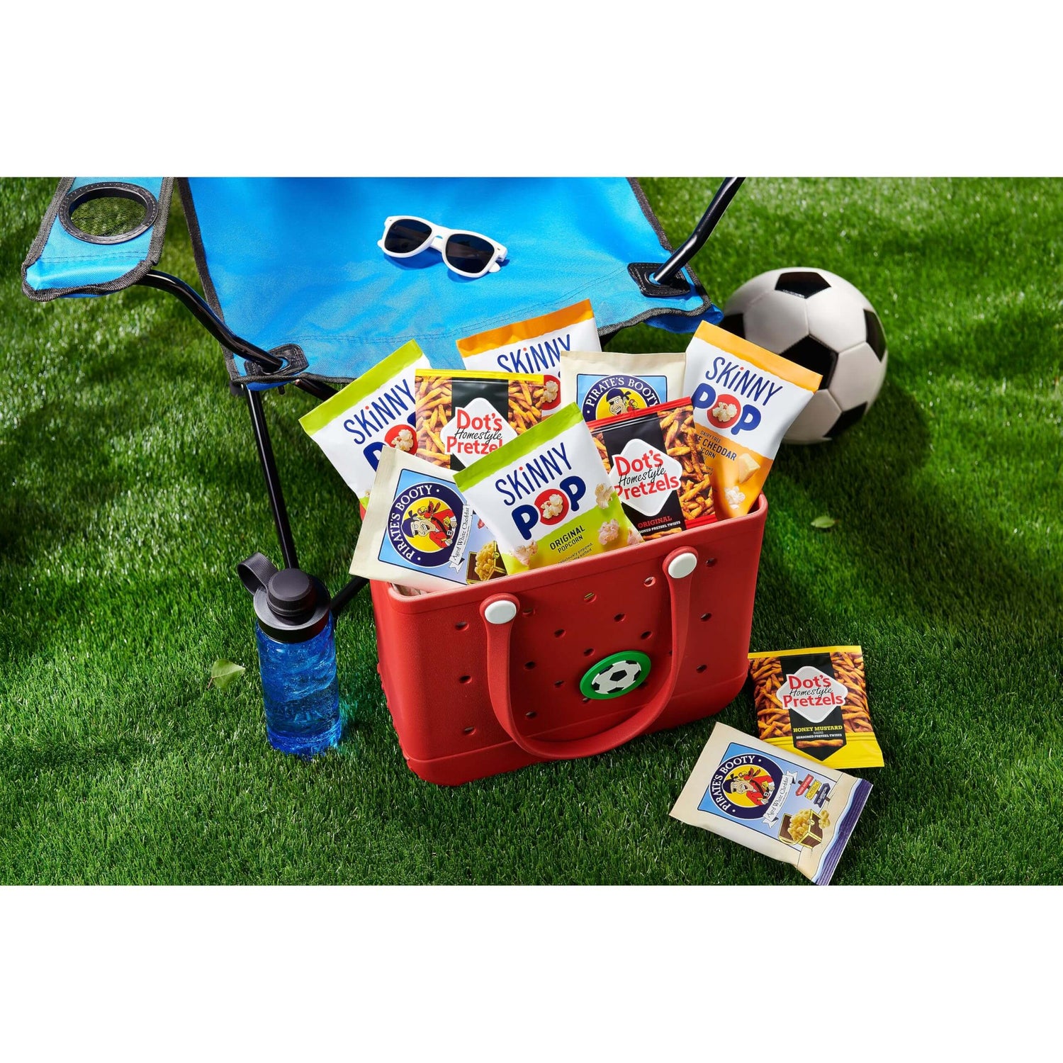 SkinnyPop Family Favorite Variety Pack snacks in a red tote bag with a soccer ball and a chair for outdoor fun.